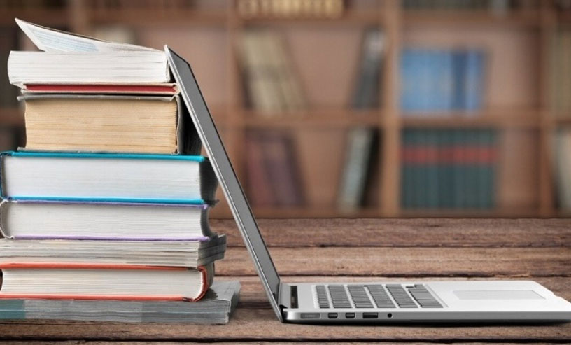 Stack of books leaning on an open laptop with a blurred bookshelf background, representing autobiography writing services.