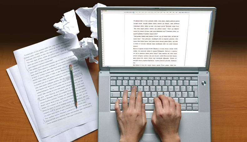 A person typing on a laptop with scattered papers and a pen nearby, representing biography writing services in action.