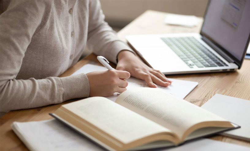 A woman writes in a notebook at her desk, laptop open, inspired by the best books on writing fiction.