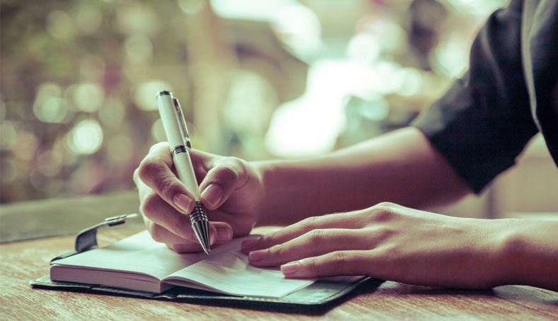 A woman writing a memoir with a pen in a notebook on a wooden table, illustrating memoir writing services.