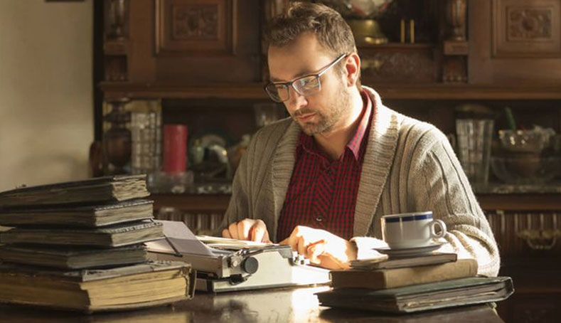 A man in glasses types on an old typewriter, focused on writing non-fiction books.