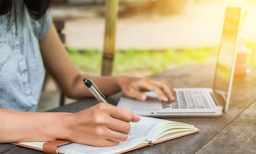 A woman focused on business writing books, simultaneously typing on a laptop and writing in a notebook.