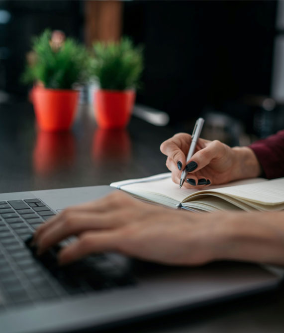 A woman engaged in writing on a notebook alongside her laptop, representing book proofreading services.