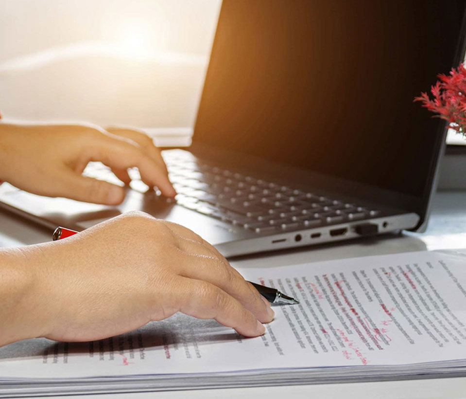 A person focused on writing with a pen on paper next to a laptop, showcasing the need for book proofreading services.